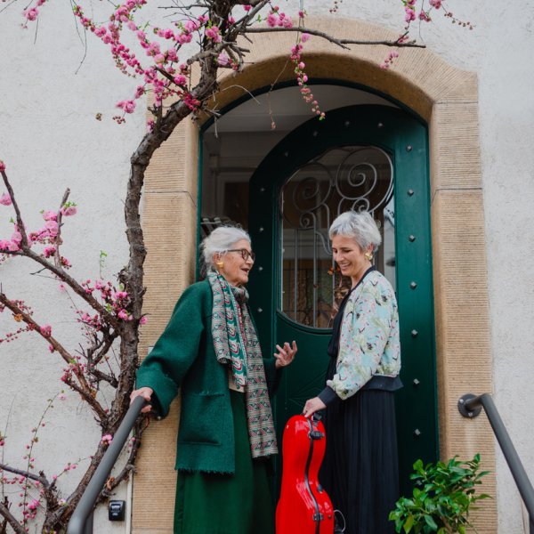 zwei Personen stehen vor einer grünen Haustür, die linke, ältere Person spricht beschwingt zur rechten Person mit einem Streichinstrumenten-Koffer, sie lacht.